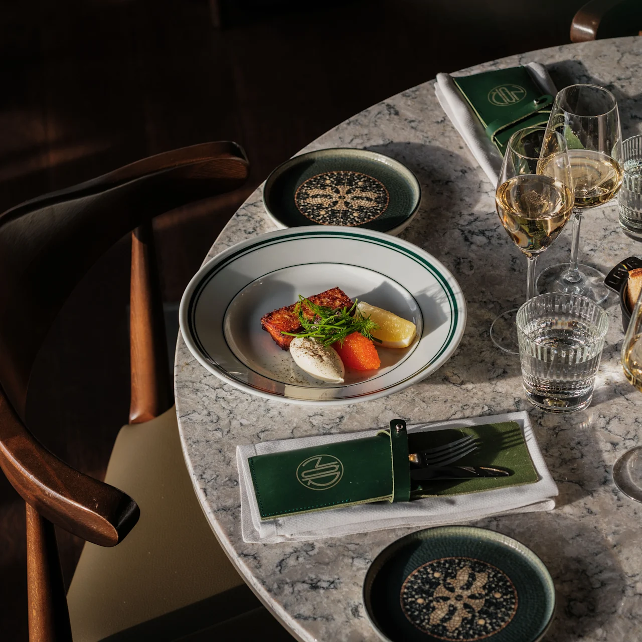 A plate of assorted cheese, tomato, and greens rests on a marble table surrounded by chairs. Glasses of white wine and a notepad with a logo are nearby.