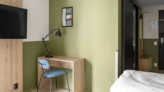 Ett träbord med en svart lampa och en blå stol står mot en grön vägg. En TV är monterad i närheten. En svart skjutdörr leder till en hall med spegel och klädhängare.