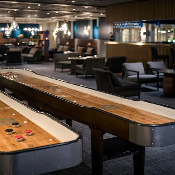 To shuffleboardbord er plassert i en moderne, dempet belyst lounge med komfortable sitteplasser og lysekroner, som skaper en avslappet og innbydende atmosfære.
