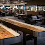 To shuffleboardbord er plassert i en moderne, dempet belyst lounge med komfortable sitteplasser og lysekroner, som skaper en avslappet og innbydende atmosfære.
