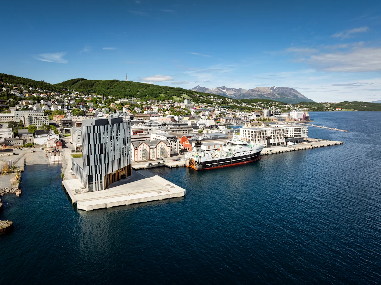 Quality Hotel Harstad, en moderne bygning på en brygge i en livlig byhavn med et stort skip fortøyd, omgitt av åser og fjell.