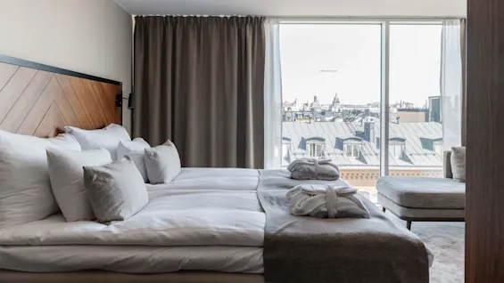 En snyggt bäddad säng framför ett stort fönster som visar en stadsvy, antyder ett modernt hotellrum.