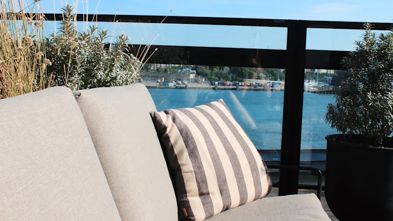 A beige outdoor couch with a striped pillow is situated on a balcony overlooking a scenic waterfront, surrounded by potted plants and clear skies.