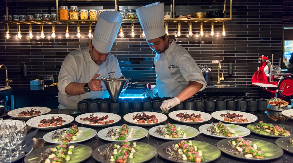 Kaksi kokkia, korkeissa hatuissa, laittavat ruokia modernissa, hämärästi valaistussa keittiössä mustilla laatoilla. Useita lautasia on aseteltu tiskille, ja niissä on gourmet-ruokia.