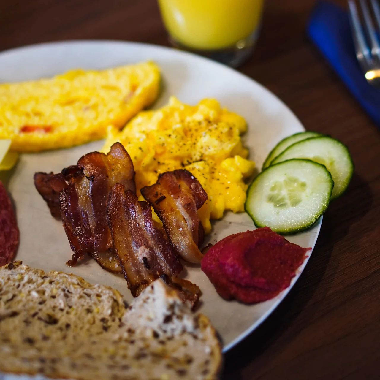 En tallerken med røræg, bacon, omelet, skiver af agurk, rødbedepuré og brød, serveret med et glas appelsinjuice på et træbord.