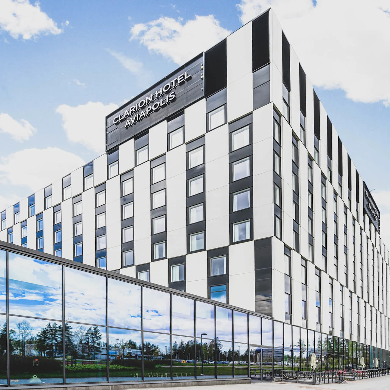 A multi-story hotel building with a checkered facade stands under a blue sky. Large windows reflect trees. Text: "CLARION HOTEL AVIAPOLIS."