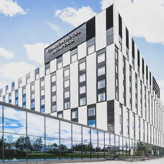 Monikerroksinen hotellirakennus ruudullisella julkisivulla seisoo sinisen taivaan alla. Suuret ikkunat heijastavat puita. Teksti: "CLARION HOTEL AVIAPOLIS."