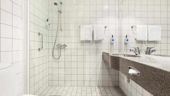 Et badeværelse har en bruser med glasdør og tilstødende dobbel vask. Flisebelagte vægge og gulv skaber et rent, moderne look, med hvide håndklæder pænt placeret på stativer.