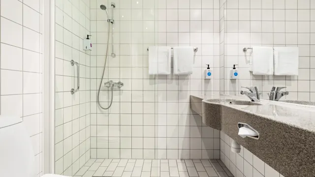 Ett badrum har en dusch med glasdörr och intilliggande dubbelhandfat. Kakel på väggar och golv ger ett rent, modernt utseende, med vita handdukar prydligt placerade på stänger.