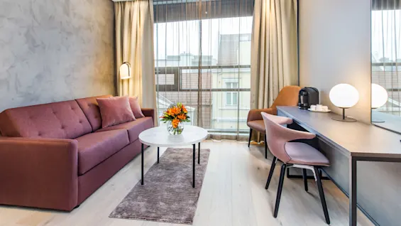 A maroon sofa sits in a modern living room beside a window. A white table with flowers is centered on a rug, and a desk with a lamp and coffee maker is nearby.