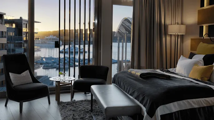 A luxurious hotel room features a bed and seating area, overlooking a marina with yachts at twilight through large windows. The decor includes neutral tones and soft lighting.