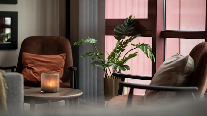 Inviting lounge area with comfortable seating, a warm candle glow, and lush greenery next to a window.