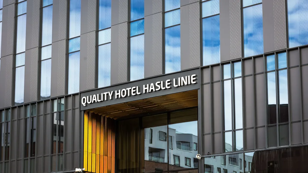 A modern hotel entrance features large glass windows reflecting blue sky and adjacent buildings. Signage reads "QUALITY HOTEL HASLE LINIE" above the entrance with a yellow accent.