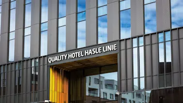 En moderne hotellinngang har store glassvinduer som gjenspeiler blå himmel og nærliggende bygninger. Skiltet lyder “QUALITY HOTEL HASLE LINIE” over inngangen med en gul aksent.