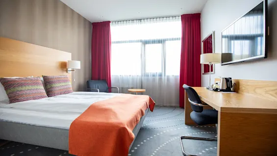 En seng med farverigt puder og et orange tæppe står i et hotelværelse med skrivebord, stol og vægmonteret TV. Røde gardiner dækker et stort vindue.