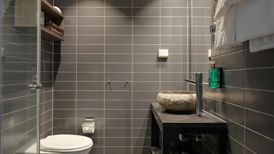 A modern bathroom features a stone sink on a black counter and a wall-mounted toilet. Grey tiles cover the walls, with a glass shower enclosure and wooden shelves.