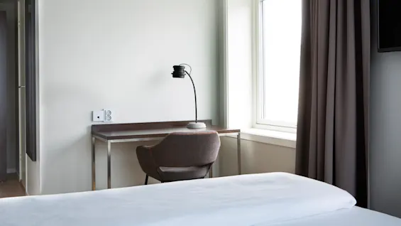 En stilren skrivebordslampe står på et trebord ved siden av en stol og et stort vindu med gardiner. Nøytrale farger, minimalistisk design.
