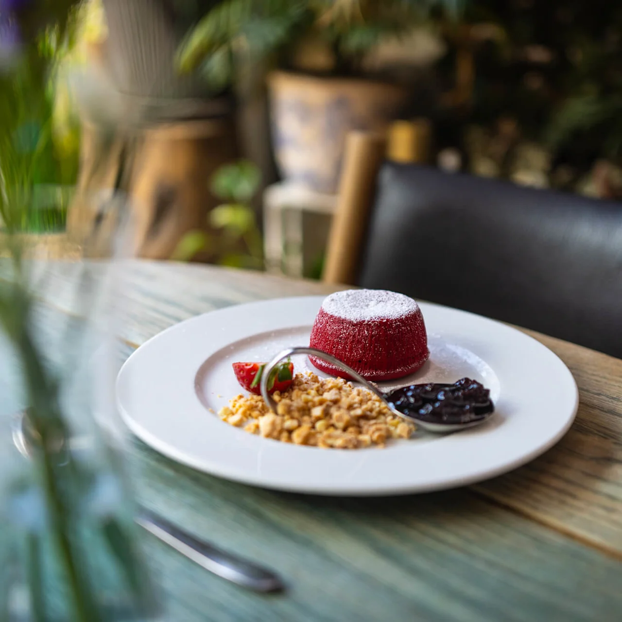 Rød kuppelformet dessert drysset med melis på en tallerken med knuste nøtter, skiver av jordbær og en skje mørk syltetøy, på et trebord i et koselig, grønt miljø.