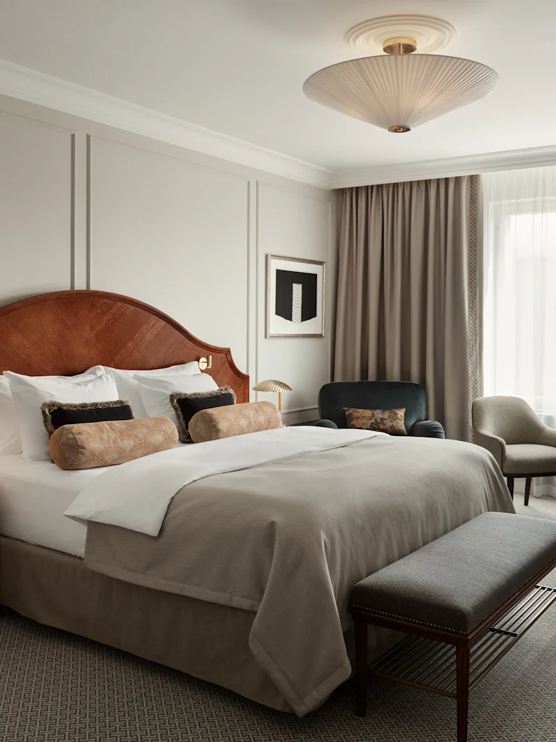 En välbäddad dubbelsäng med kuddar och en trähuvudgavel står i ett hotellrum. Bredvid finns ett bord med två stolar, ett fönster med gardiner och en stilfull taklampa.