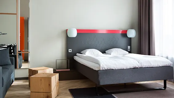 Et moderne hotellrom har en pent oppredd dobbeltseng med hvitt sengetøy, et geometrisk hodegjerde og dekorative puter. Trestoler og et speil gir stil til det lyse rommet.