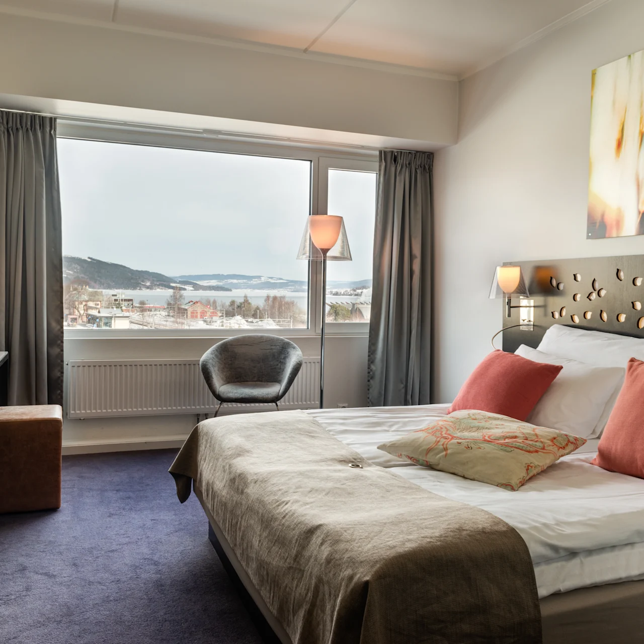 A hotel room features a bed with decorative pillows, a desk with a chair, and a large window showing a scenic view of hills and water.