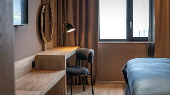 Et træskrivebord med et rundt spejl og en tændt lampe står ved siden af et vindue med brune gardiner. En stol står ved skrivebordet, og en del af en seng kan ses.