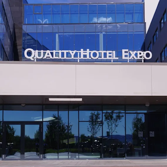 Fasade med store vinduer, viser skiltet "QUALITY HOTEL EXPO" over inngangen. Moderne arkitektur, omgitt av trær og blå himmel reflektert.