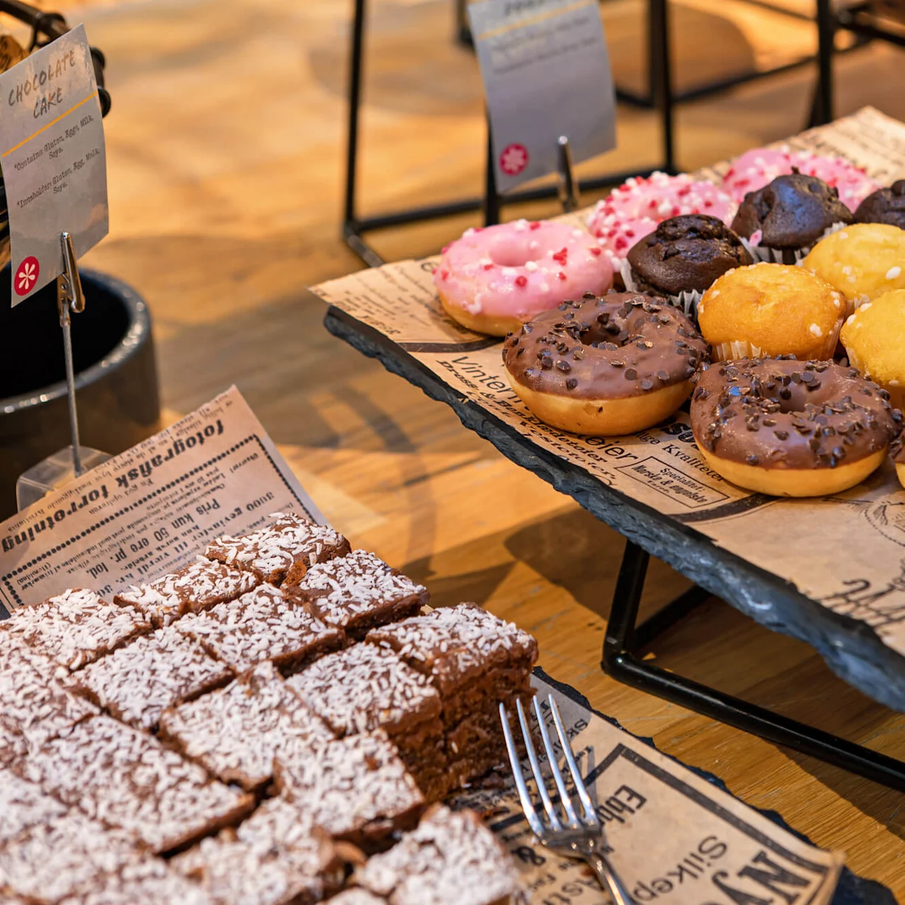 Assorterte donuts og muffins ligger på et skiferbrett, omgitt av avisstilark. I nærheten er firkantede sjokoladekaker toppet med kokos. Tekst: "Chocolate Cake" og "Holgeristik forretning".