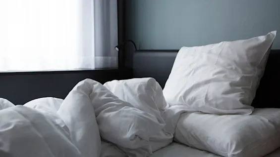 Seng med krøllede hvide lagner og puder, ved siden af et vindue med tynde gardiner. Blød belysning skaber en rolig, minimalistisk soveværelsesstemning.
