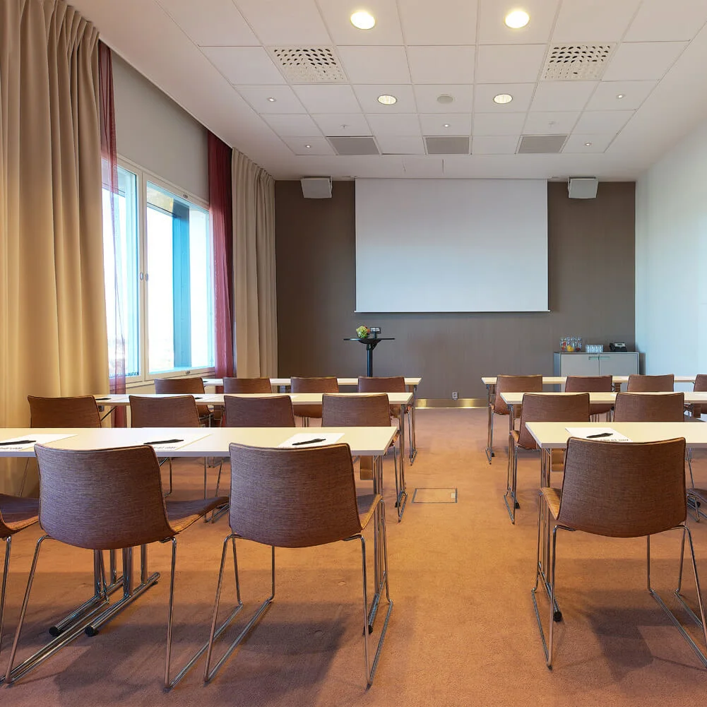 A conference room with arranged tables and chairs facing a large screen. Soft lighting illuminates the space, with curtains on the windows and a cabinet with drinks at the back.