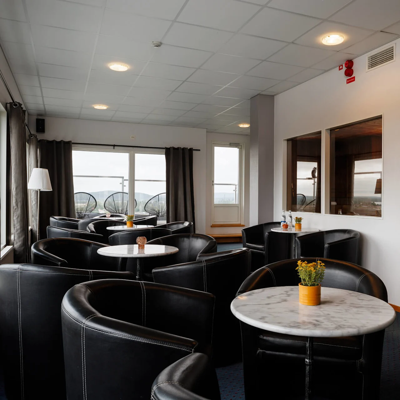 Bekväma svarta läderstolar omger marmorbord prydda med små växter, belägna i en lounge nära en glasinramad bastu; stora fönster avslöjar en utsikt över en balkong utomhus.