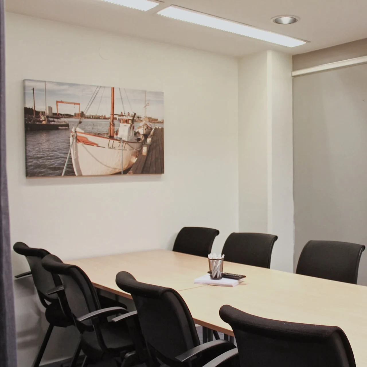 Conference table surrounded by black chairs, holding a notepad and pen cup. Walls display boat painting and a blank flip chart. Fluorescent lighting overhead.