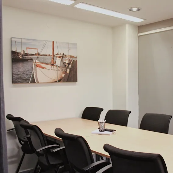Konferensbord omgivet av svarta stolar, med en anteckningsblock och pennkopp. Väggar pryds av båtbild och en tom blädderblock. Lysrörsbelysning ovanför.