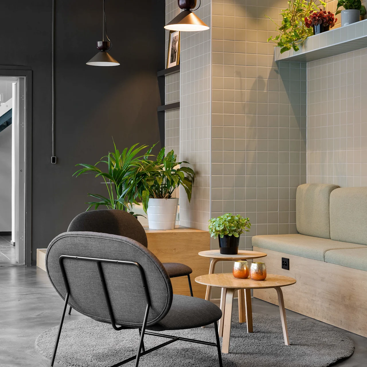 A modern lounge features a gray chair facing a small wooden table with candles, surrounded by plants. Overhead, pendant lights illuminate the tiled wall setting.
