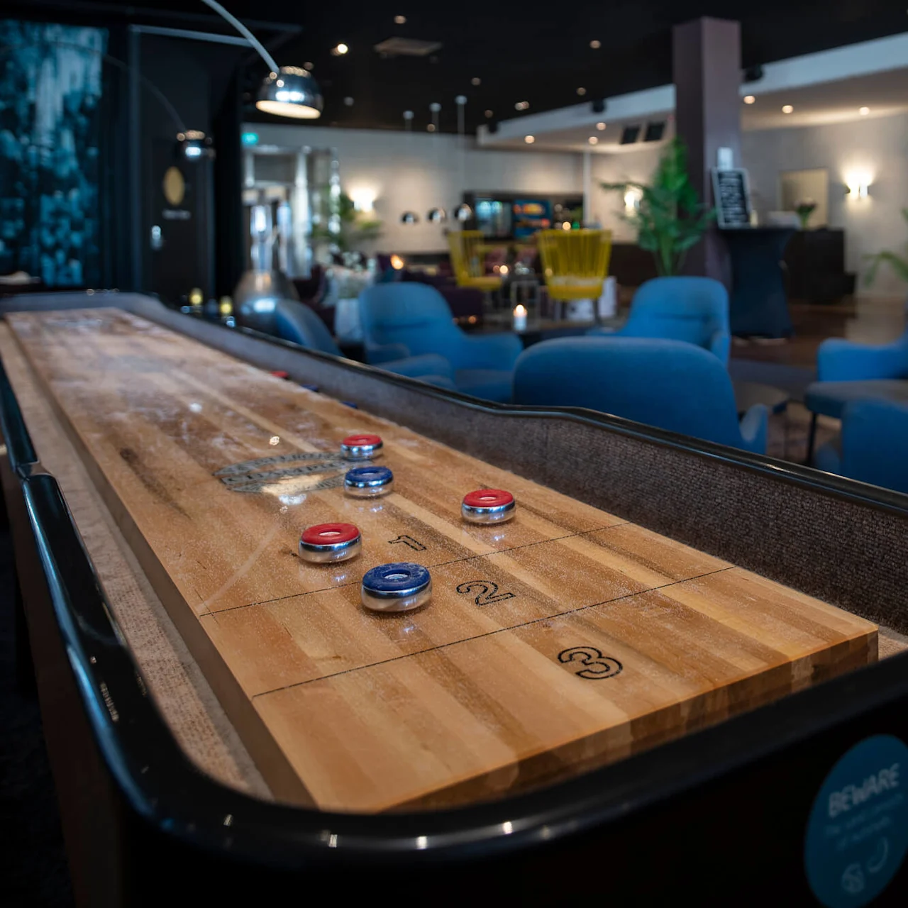 En shuffleboardbord med röda och blå pjäser står i en mysig loungemiljö med blå stolar, växter och hyllor. Text: "BEWARE Mind your fingers."