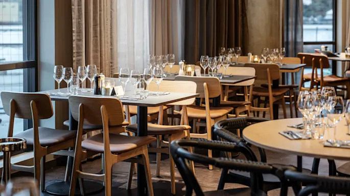 Tables set with wine glasses and napkins stand in an elegant restaurant dining area with light wooden chairs, surrounded by large windows and soft, flowing curtains.