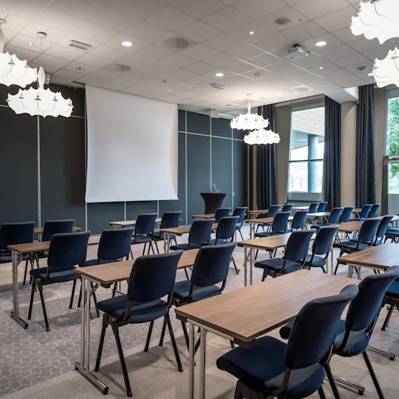 Stoler og bord er arrangert i rader mot en stor projektor skjerm i et konferanserom. Elegante lysekroner henger fra taket, med store vinduer som gir naturlig lys.