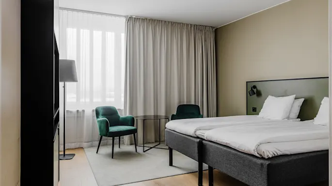 En pent oppredd seng med hvitt sengetøy er i et moderne soverom. To grønne stoler og et lite bord er i nærheten, under et høyt vindu med gardiner.