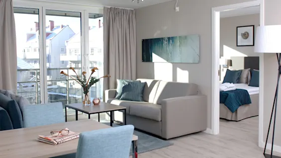 Stue med beige sofa og turkise puter, foran et spisebord med avis og briller. Store vinduer med utsikt til nabobygninger, koblet til soverom med turkist sengetøy.