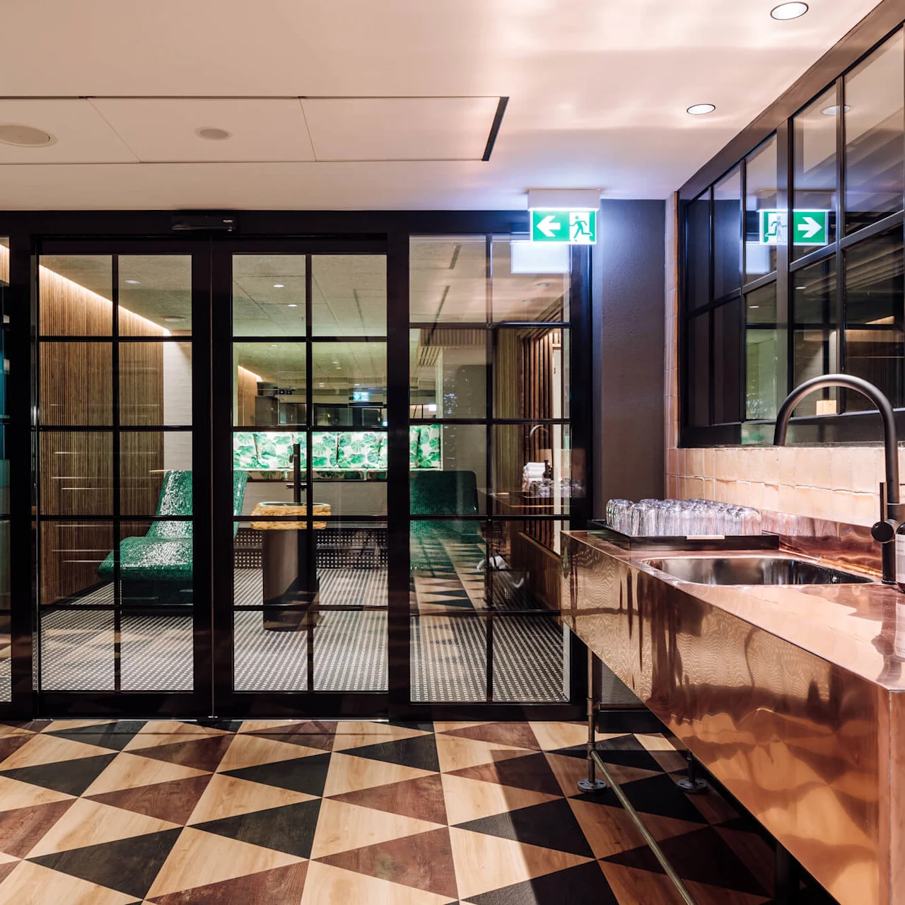 Sink holds stacked towels and soap, adjacent to glasses; glass doors open to seating area with green chairs, geometric patterned floor. Exit signs with running figure and arrow overhead.
