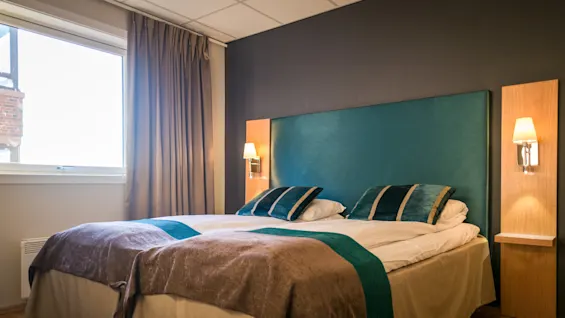 A neatly made hotel bed with teal and brown accents, flanked by wall lamps, stands in a room with wooden flooring and beige curtains covering a large window.