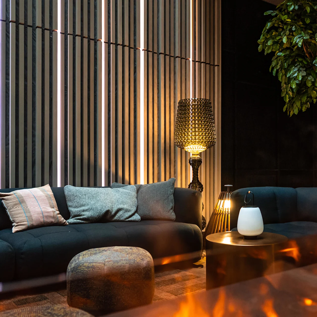 En modern lounge-sektionsplats har en mörk soffa med kuddar, en tänd dekorativ lampa och en krukväxt mot en randig vägg; flammor fladdrar i förgrunden.