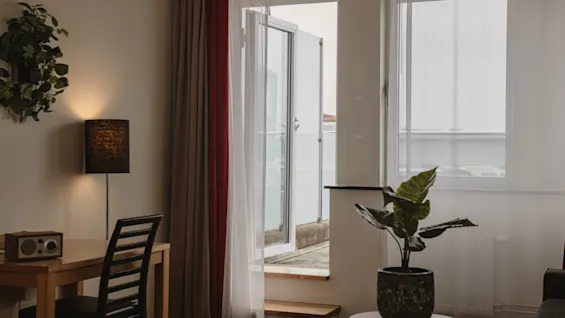 A chair stands next to a lamp and table by a doorway. Sheer curtains frame an open door leading to a balcony. A plant sits on a round table nearby.