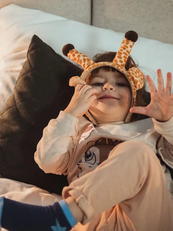 Lapsi, jolla on kirahvipanta, makaa leikkisästi sängyllä, hymyilee ja nostaa kätensä.