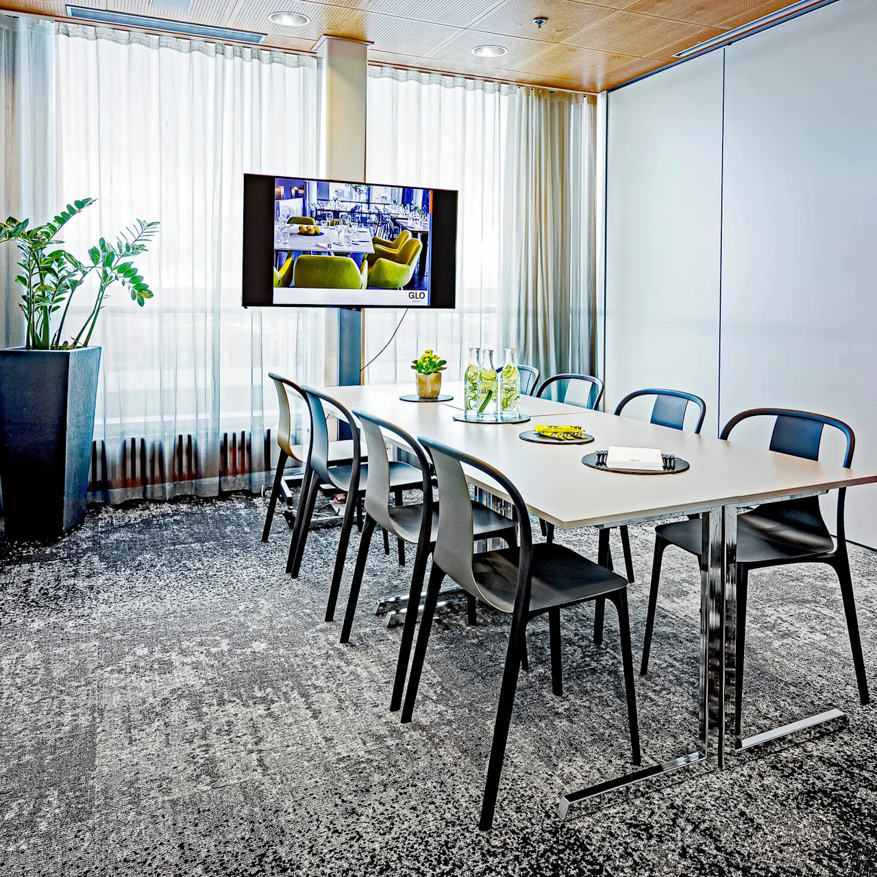 Conference room table with black chairs arranged around it, facing a mounted screen displaying "GLO". A potted plant stands by a window with sheer curtains.