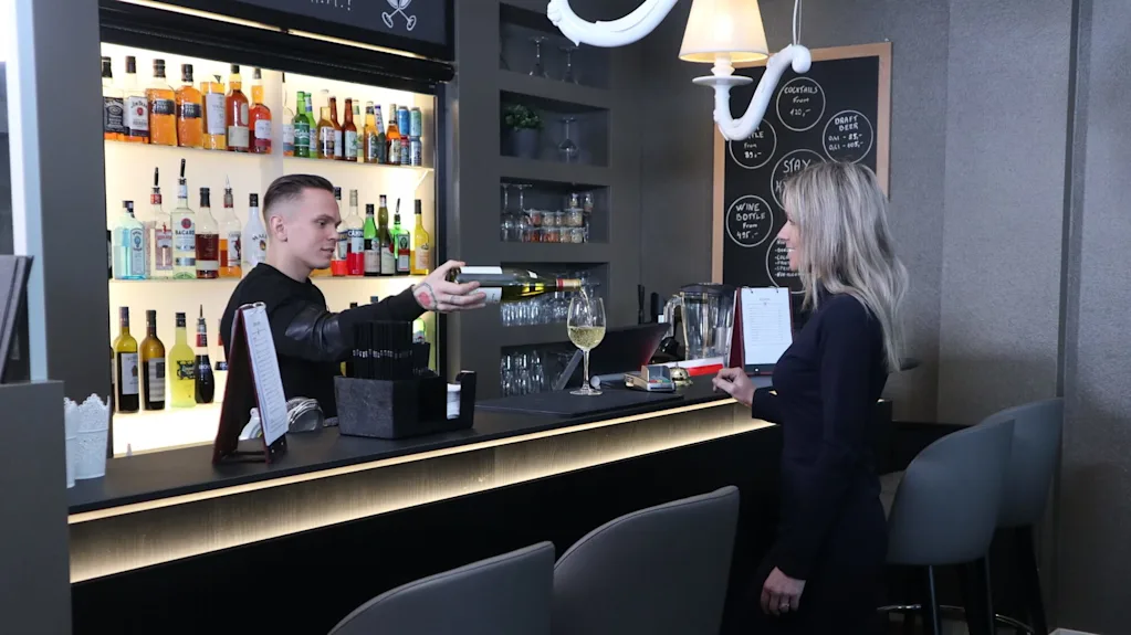 En bartender rækker et kort til en kunde ved en moderne bar med hylder af flasker og en tavle der viser drikkevarer.