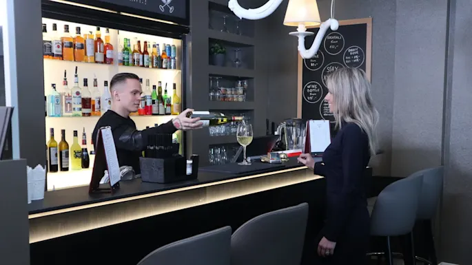 A bartender is handing a card to a customer at a modern bar with shelves of bottles and a blackboard listing drinks.