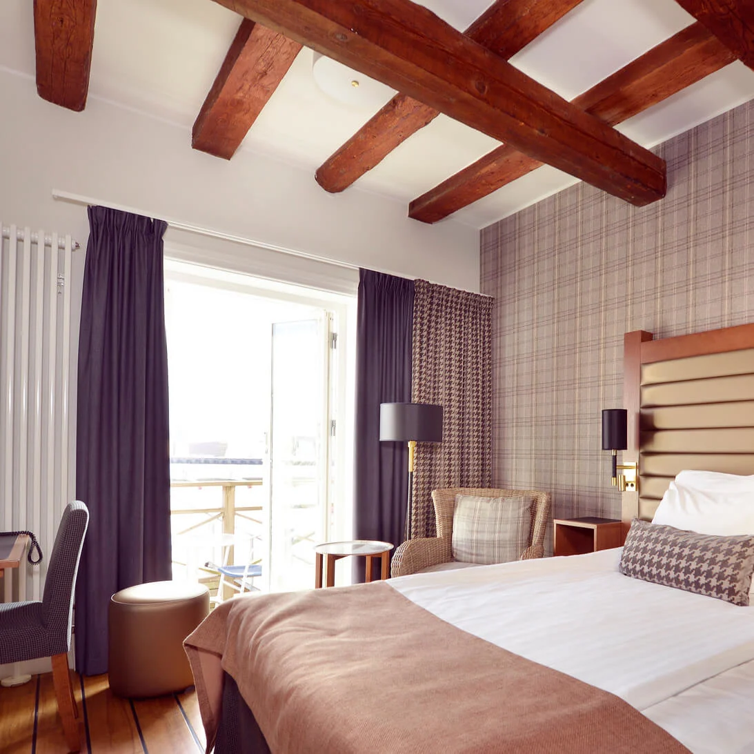A tidy bedroom with a made bed, wooden desk, and chair, set in a room with wooden ceiling beams and a balcony.