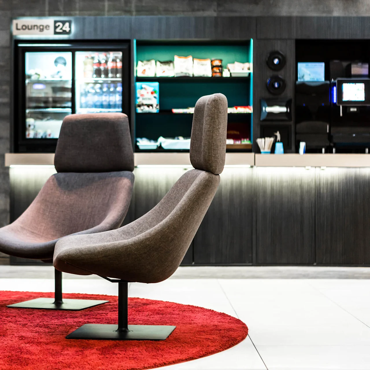 Gray lounge chairs sit on a red carpet in front of a snack bar with a refrigerator and shelves displaying snacks and drinks. Text: "Lounge 24".