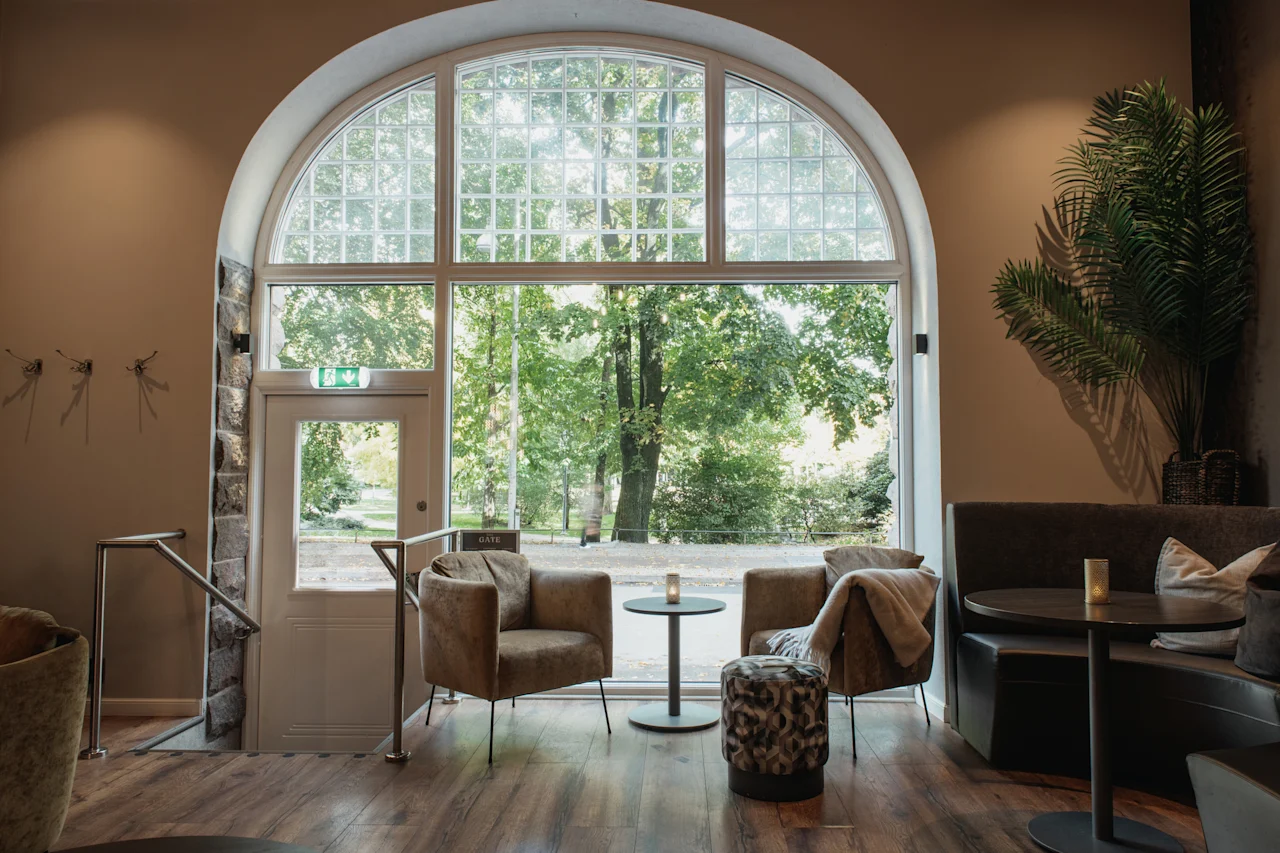 Et hyggeligt loungeområde med komfortable siddepladser og store buede vinduer med udsigt over frodige grønne træer. En dør med teksten GATE er synlig.